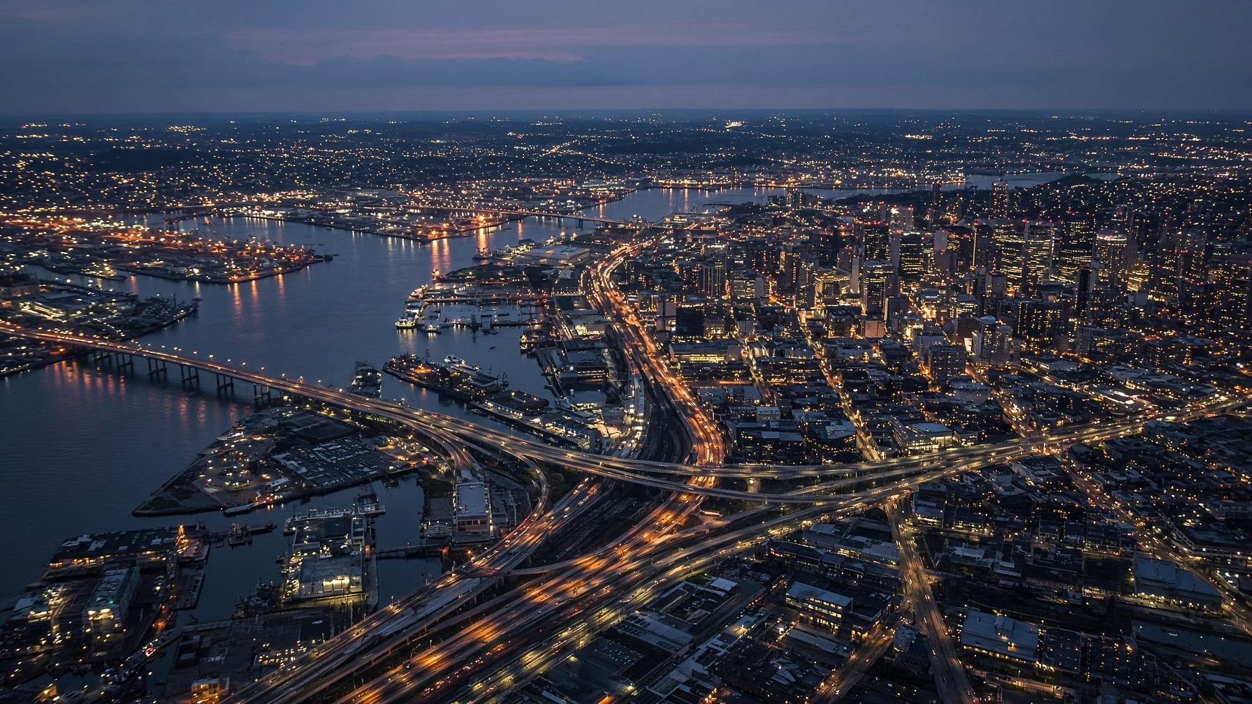 Aerial of a harbor city at blue hour, light moving through streets and water.